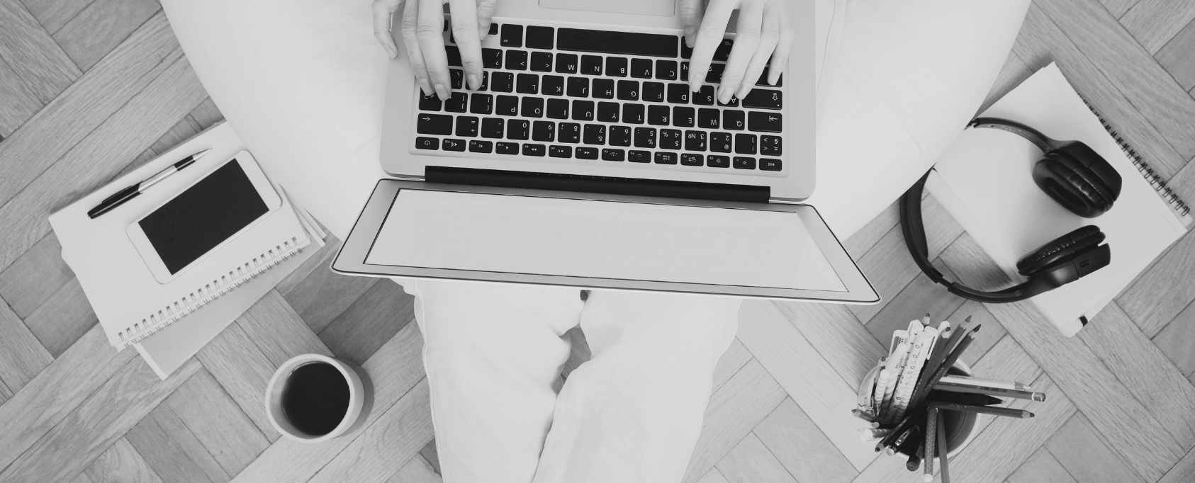 Someone sitting in a chair with laptop on their lap. Coffee, headphones, notepad and pencils are on the floor.